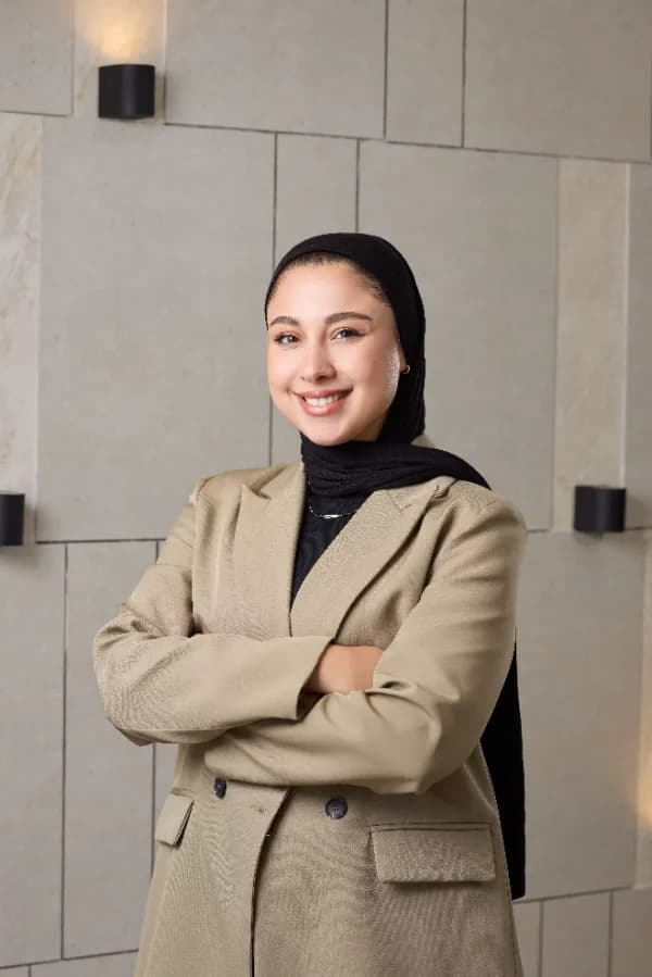 Woman in a beige blazer and black headscarf standing with arms crossed