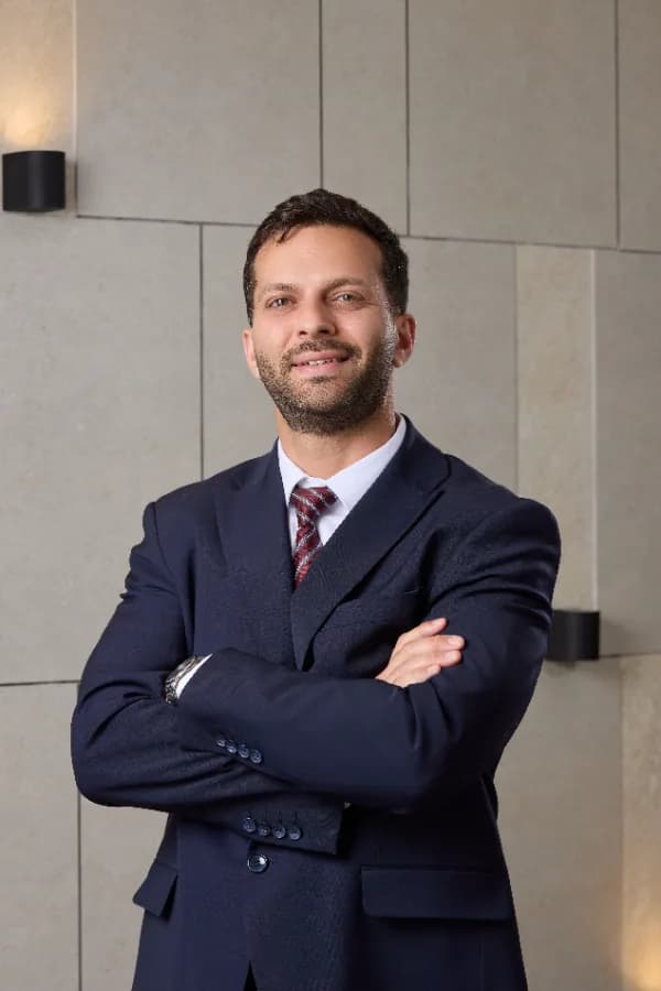 Man in a navy suit standing with arms crossed against a stone-tiled wall