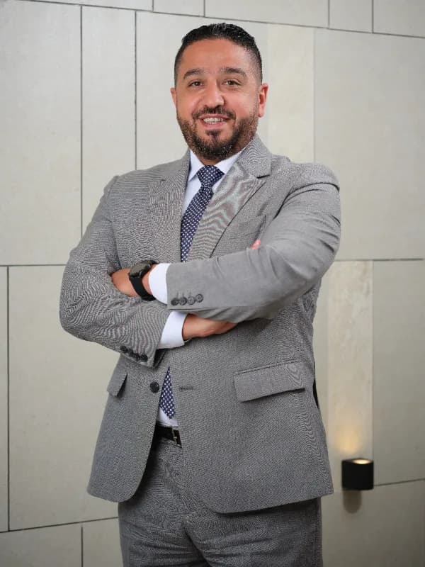 Man in a grey suit standing with arms crossed against a stone-tiled wall