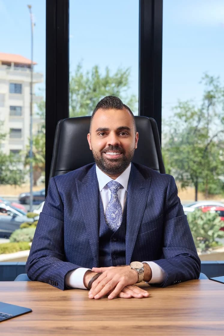 Rami Musallam in a blue suit, seated at a desk in front of floor-to-ceiling office windows